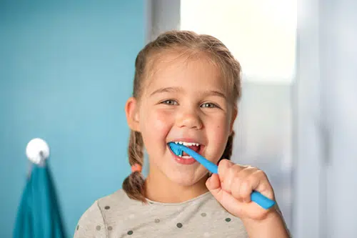 Young girl brushing teeth with blue toothbrush in bathroom with blue towel.