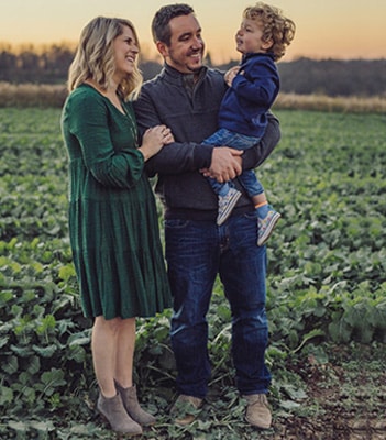Family of three standing in a soybean field at sunset, smiling together outdoors.