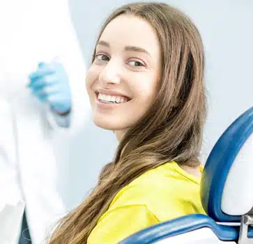Smiling young woman in yellow shirt at dental office with blue chair visible.