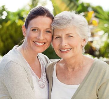 Two women smiling outdoors with green foliage background, representing family dental care.