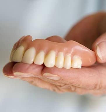 Hand holding upper denture teeth showing dental prosthetic restoration against neutral background.