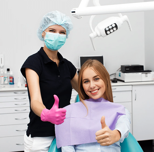 Dental hygienist in surgical mask and cap gives thumbs up with young patient in treatment room.