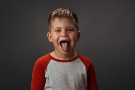 Young boy in red and gray shirt sticking tongue out playfully against gray background.