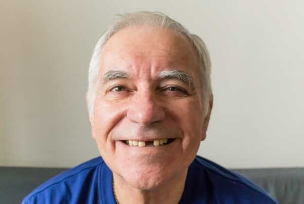 Smiling older man with white hair wearing blue shirt against neutral background at dental office.