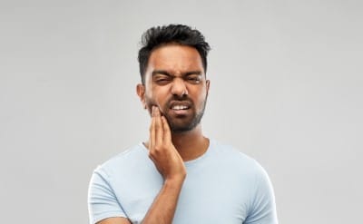 Man with glasses touching jaw in pain against gray background, dental discomfort.
