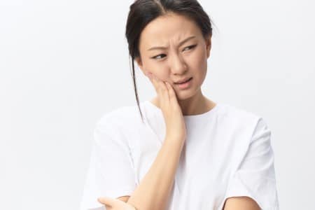 Woman in white shirt touching face thoughtfully against white background, dental care concept.