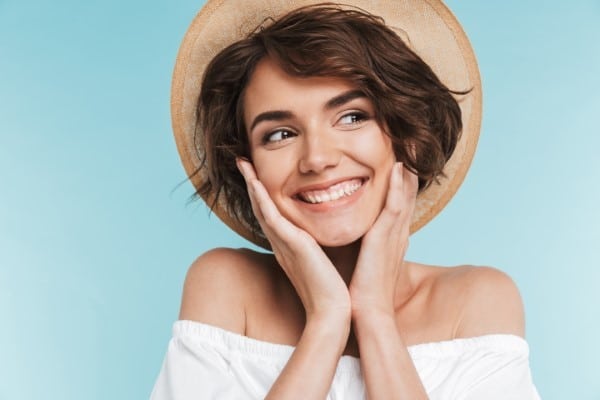 Smiling woman wearing straw hat and white off-shoulder top against blue background, touching face with hands.
