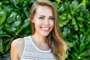 Smiling woman in light tank top posed outdoors against green foliage background.