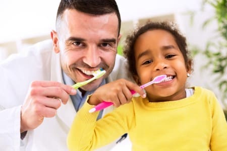 Father and young child brushing teeth together in bright, clean dental office setting.