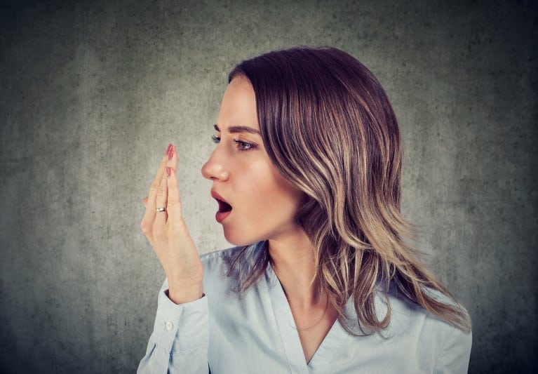 Woman checking breath with hand near mouth against neutral background.