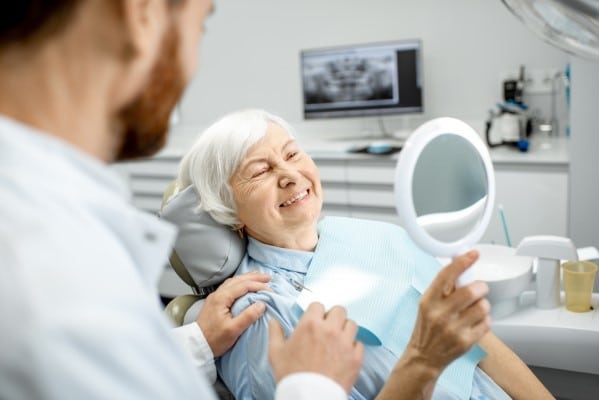 Senior woman examines face in mirror during dental appointment with dentist in modern clinic.