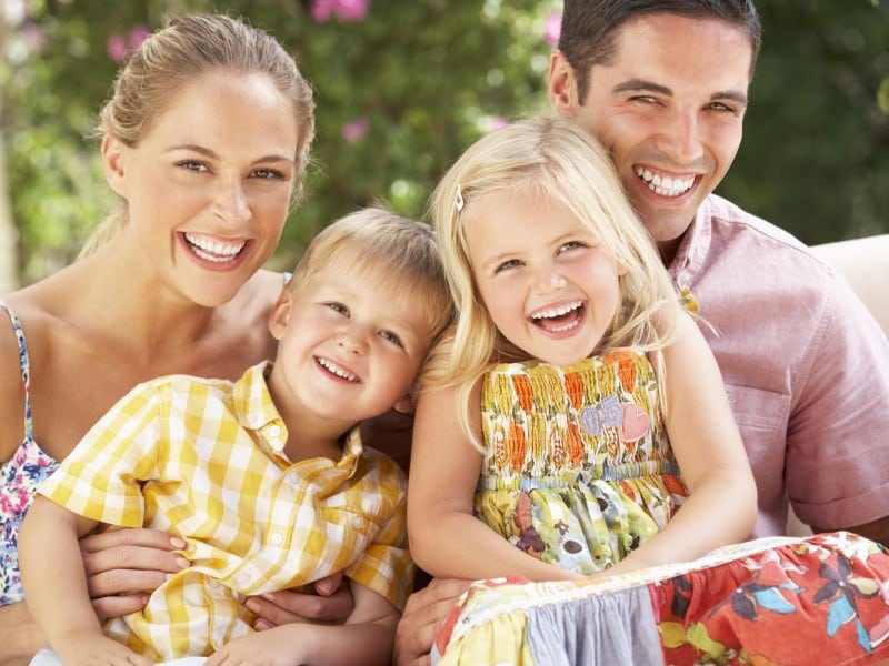 Happy family of four smiling outdoors in casual clothing, enjoying time together in garden setting.