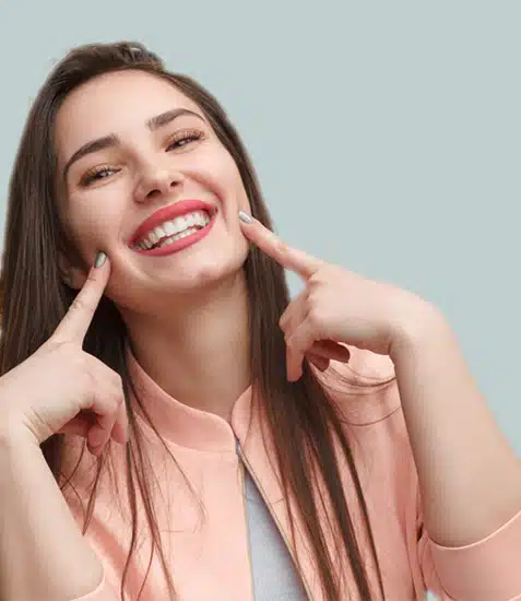 Woman smiling brightly while pointing to her teeth against neutral gray background.