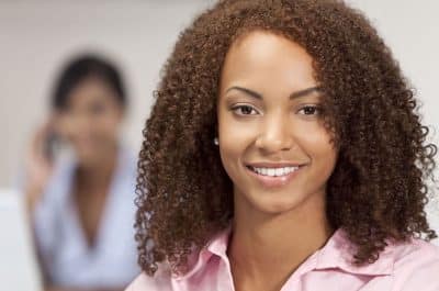 Smiling woman in pink shirt at dental office with colleague blurred in background.