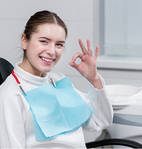 Young female dental patient smiling and giving okay sign in dental office treatment room.