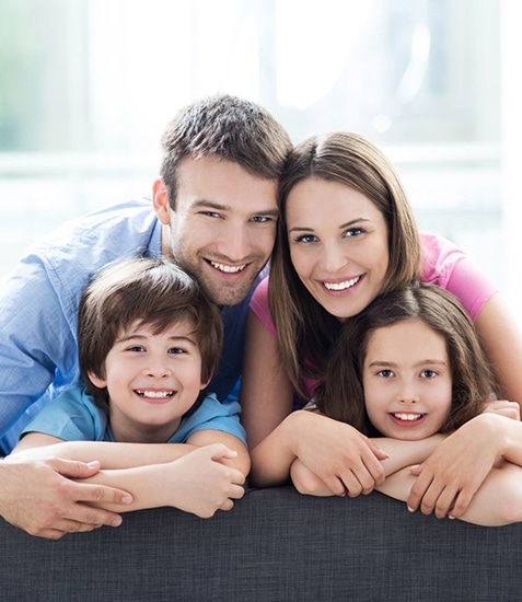 Happy family of four smiling together indoors near bright window at dental office.